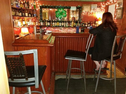 A cozy bar scene with a woman sitting on a stool, surrounded by bottles and festive decorations.