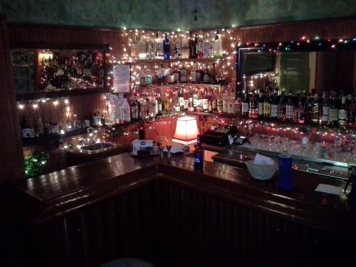 A cozy bar with shelves of liquor bottles, decorated with colorful lights and festive garlands.