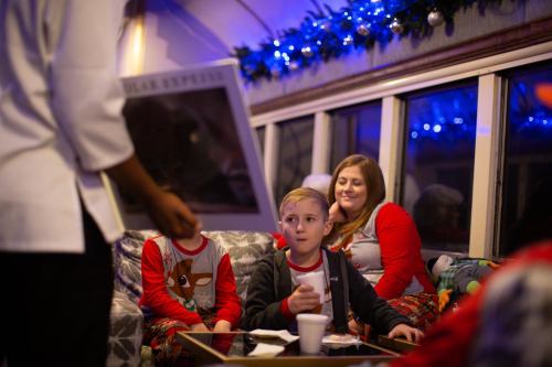A child in festive pajamas sits at a table, looking surprised, while an adult holds up a poster in a cozy, decorated setting.
