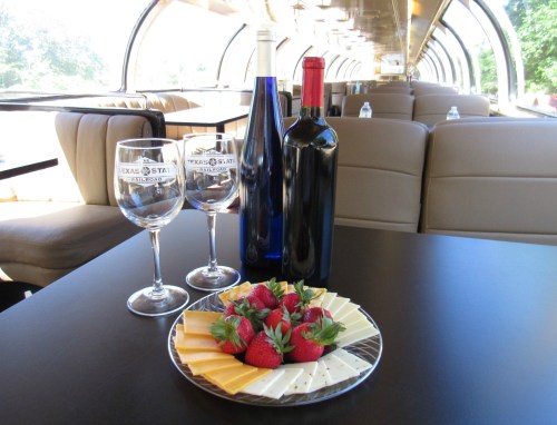 A table set with two wine glasses, three bottles of wine, and a plate of strawberries and cheese in a train car.
