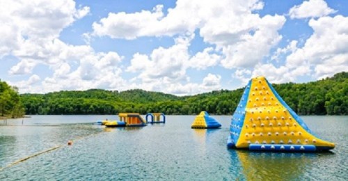 Colorful inflatable water structures on a lake, surrounded by green trees and a bright blue sky with fluffy clouds.