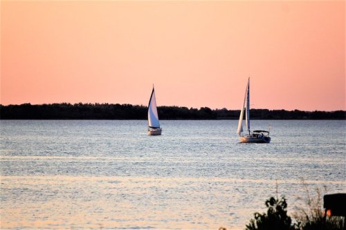 Two sailboats glide across a calm body of water at sunset, with a soft pink and orange sky in the background.