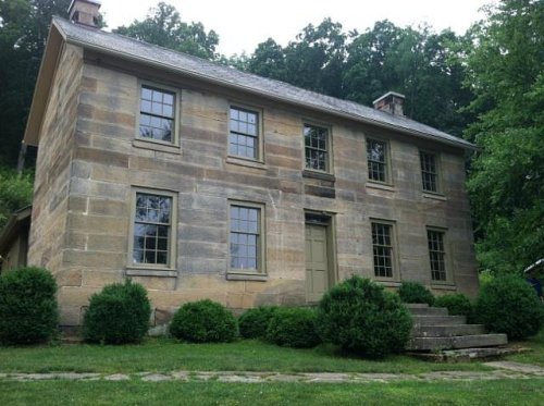 A historic stone house with large windows, surrounded by greenery and steps leading to the entrance.