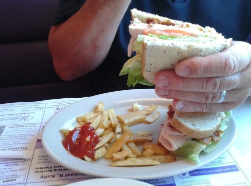 A hand holding a large sandwich with lettuce and tomato, next to a plate of fries and ketchup.