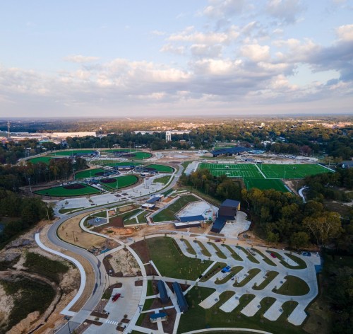 Aerial view of a large sports complex with multiple fields, pathways, and green spaces under a cloudy sky.