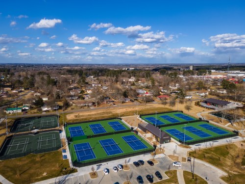 Aerial view of a tennis complex with multiple courts, surrounded by trees and a small town in the background.