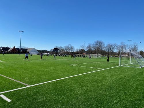 A sunny day at a soccer field with players engaged in a game on artificial turf.