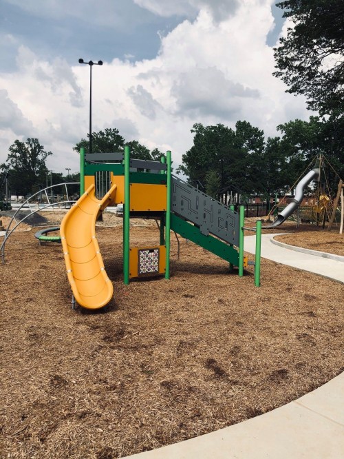 Colorful playground equipment with a yellow slide, surrounded by mulch and trees under a cloudy sky.