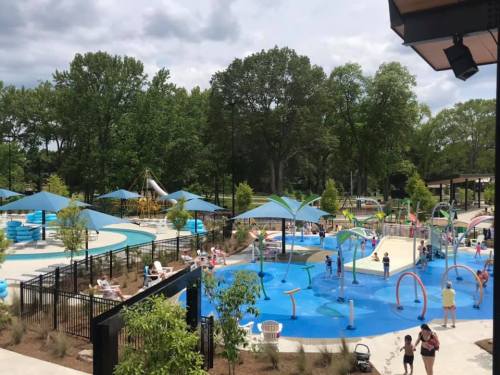A vibrant water park with slides, splash pads, and families enjoying the sunny day among green trees and blue skies.