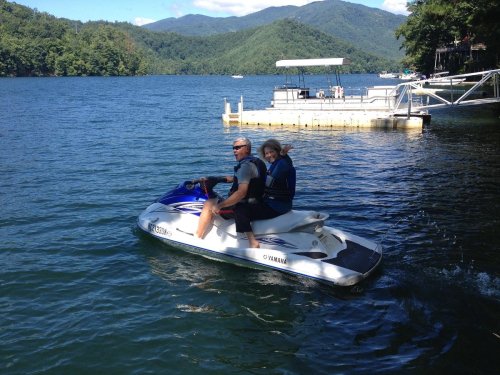 A man and woman ride a jet ski on a lake, with mountains and a dock in the background.