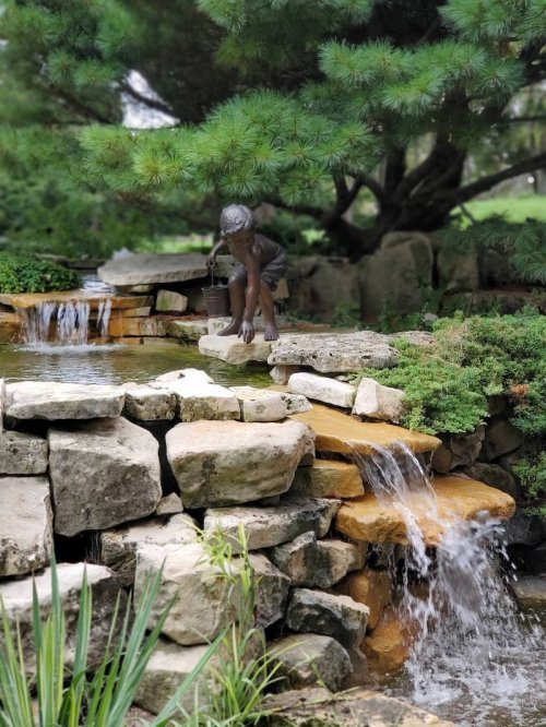 A serene garden scene featuring a stone waterfall and a statue of a child playing near the water. Lush greenery surrounds.