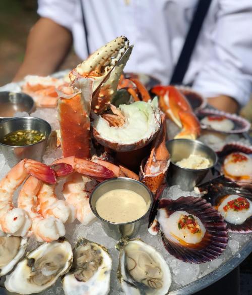 A seafood platter featuring shrimp, crab, oysters, and scallops served on ice with various sauces in small cups.