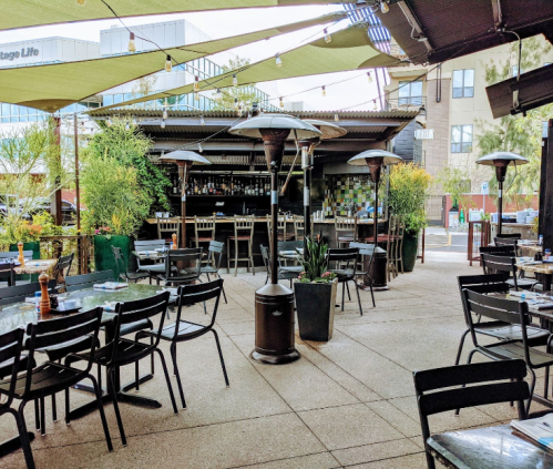 Outdoor restaurant patio with tables, chairs, and patio heaters, surrounded by greenery and a bar in the background.