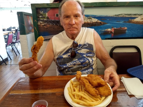 A man in a sleeveless shirt holds a piece of fried chicken, sitting at a table with a plate of chicken and fries.