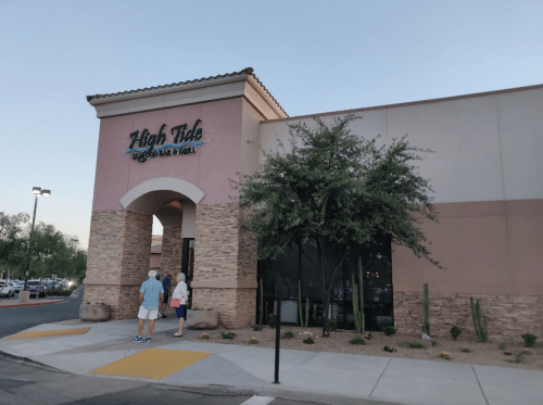 Exterior of High Tide Seafood Bar & Grill with two people standing outside, surrounded by desert landscaping.