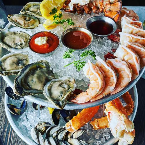 A seafood platter featuring shrimp, oysters, crab legs, and sauces on ice, garnished with lemon and parsley.