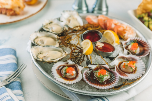 A seafood platter featuring oysters, scallops, shrimp, and lemon wedges on ice, served with dipping sauces.