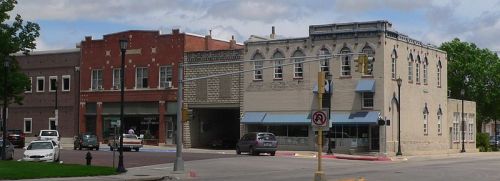 Historic buildings line a street corner, featuring brick facades and storefronts under a partly cloudy sky.