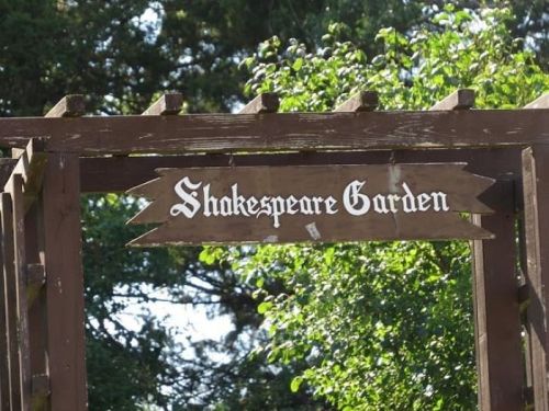 Wooden sign reading "Shakespeare Garden" hanging from a trellis, surrounded by greenery.