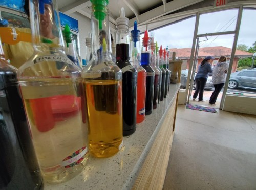 A row of colorful syrup bottles on a counter, with people visible through the doorway in the background.