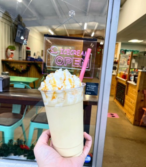 A hand holds a creamy milkshake topped with whipped cream and caramel, with an "Ice Cream Open" sign in the background.