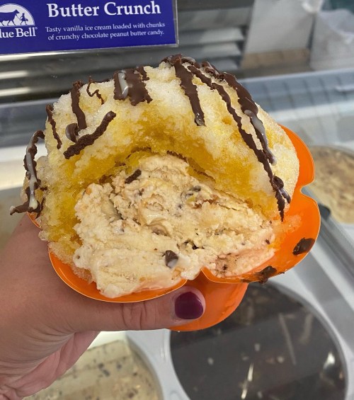 A hand holding a vibrant orange bowl filled with yellow shaved ice and vanilla ice cream drizzled with chocolate.
