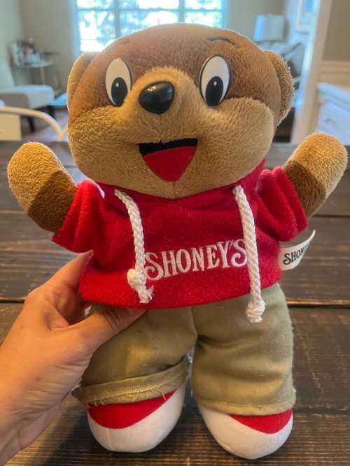 A plush bear toy wearing a red Shoney's hoodie and tan pants, smiling and standing on a wooden table.