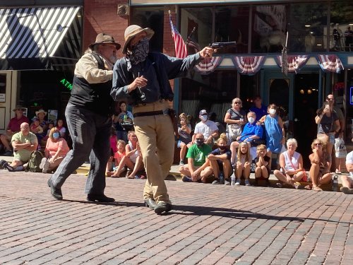 Two men in Western attire perform a staged gunfight in front of a crowd on a brick street during a festival.