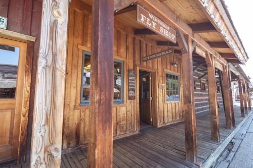A rustic wooden storefront with a sign reading "Fly Shop" and an open door, set in a charming outdoor setting.