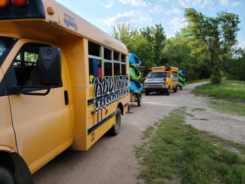 Two yellow buses parked on a dirt road, each towing kayaks, surrounded by trees and blue sky.