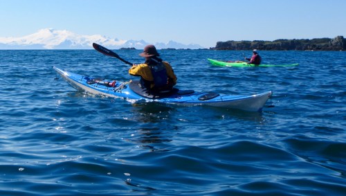 Two kayakers paddle in calm blue waters with snow-capped mountains in the background on a clear day.