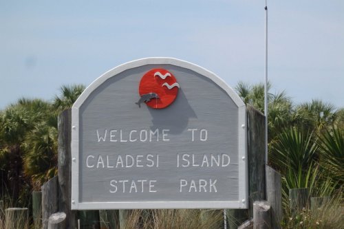 Welcome sign for Caladesi Island State Park featuring a dolphin and waves design. Lush greenery in the background.
