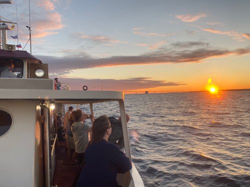 People on a boat watch a vibrant sunset over the water, with a bridge visible in the background.