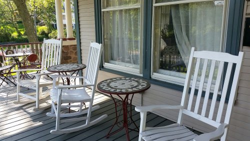 A porch with two white rocking chairs and a small round table, surrounded by greenery and sunlight.