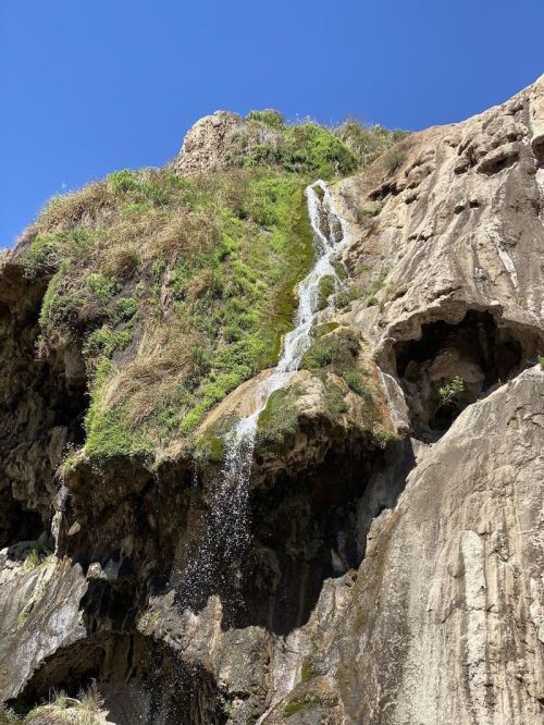 A waterfall cascading down a rocky cliff, surrounded by lush greenery under a clear blue sky.