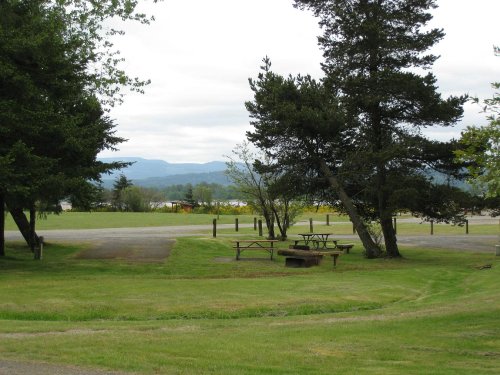 A grassy area with picnic tables, trees, and distant mountains under a cloudy sky.