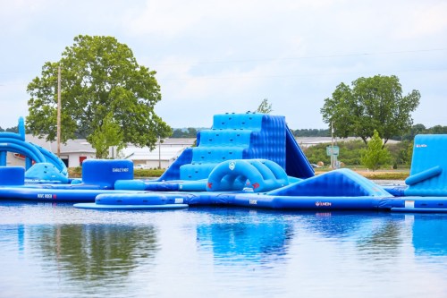 A blue inflatable water park with various structures on a calm lake, surrounded by trees and a cloudy sky.