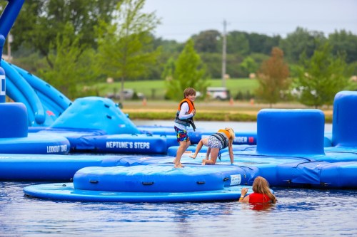 Children playing on inflatable water structures in a lake, with one child climbing and others swimming nearby.
