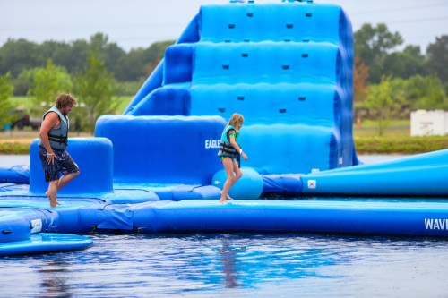 A man and a girl walk on a blue inflatable water obstacle course in a lake, surrounded by greenery.