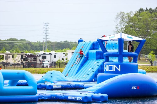 A colorful inflatable water park with a climbing wall and slides, set on a lake with trees and campers in the background.