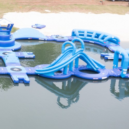 A blue inflatable water park with slides and obstacles on a lake, surrounded by grass and a sandy area.