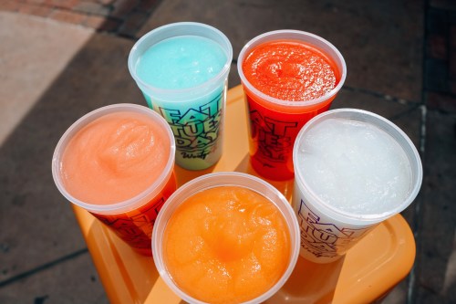 Six colorful frozen drinks in clear cups, arranged on a table, featuring shades of blue, orange, red, and white.