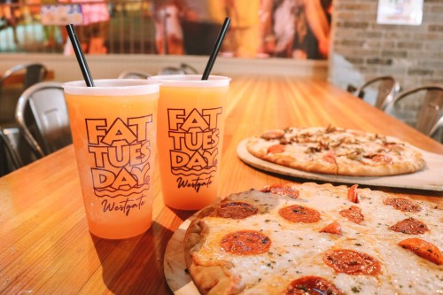 Two drinks in "Fat Tuesday" cups beside a pepperoni pizza on a wooden table. A second pizza is in the background.