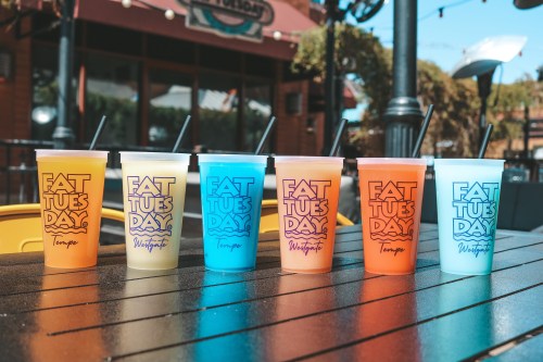 Six colorful cups with "Fat Tuesday" branding, filled with drinks, lined up on a table outdoors.