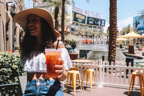 A smiling woman in a straw hat holds a drink, standing in a sunny outdoor area with palm trees and a fountain.