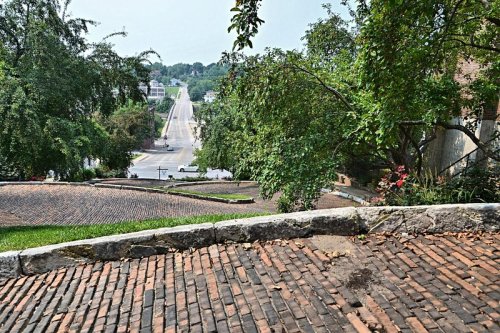 A view of a winding road from a hillside, lined with trees and brick pathways, under a clear sky.