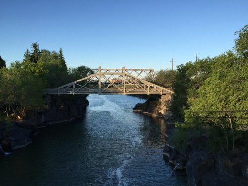 A metal bridge spans a calm river, surrounded by lush greenery and clear blue skies.