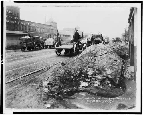 Black and white photo of a snowy street with horse-drawn wagons and blocked traffic in an urban setting.