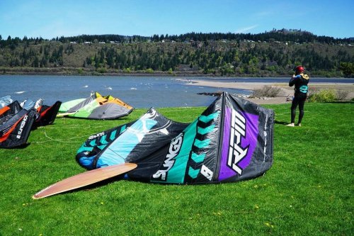 A person prepares a kite on green grass by a river, with colorful kites and a scenic landscape in the background.
