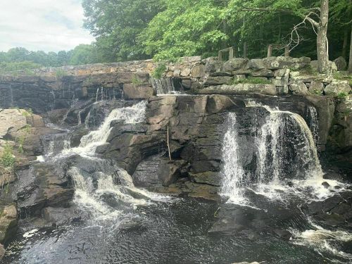 A serene waterfall cascading over rocky terrain, surrounded by lush green trees and a cloudy sky.